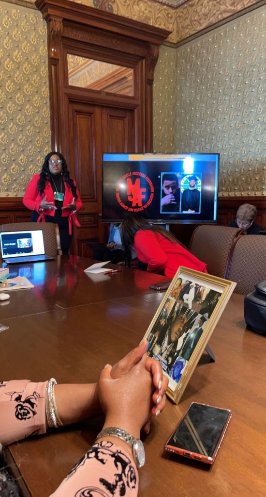 An African-American woman in a red jacket presenting at a screen at a table. The screen displays the MJAF logo and pictures of Melquain.